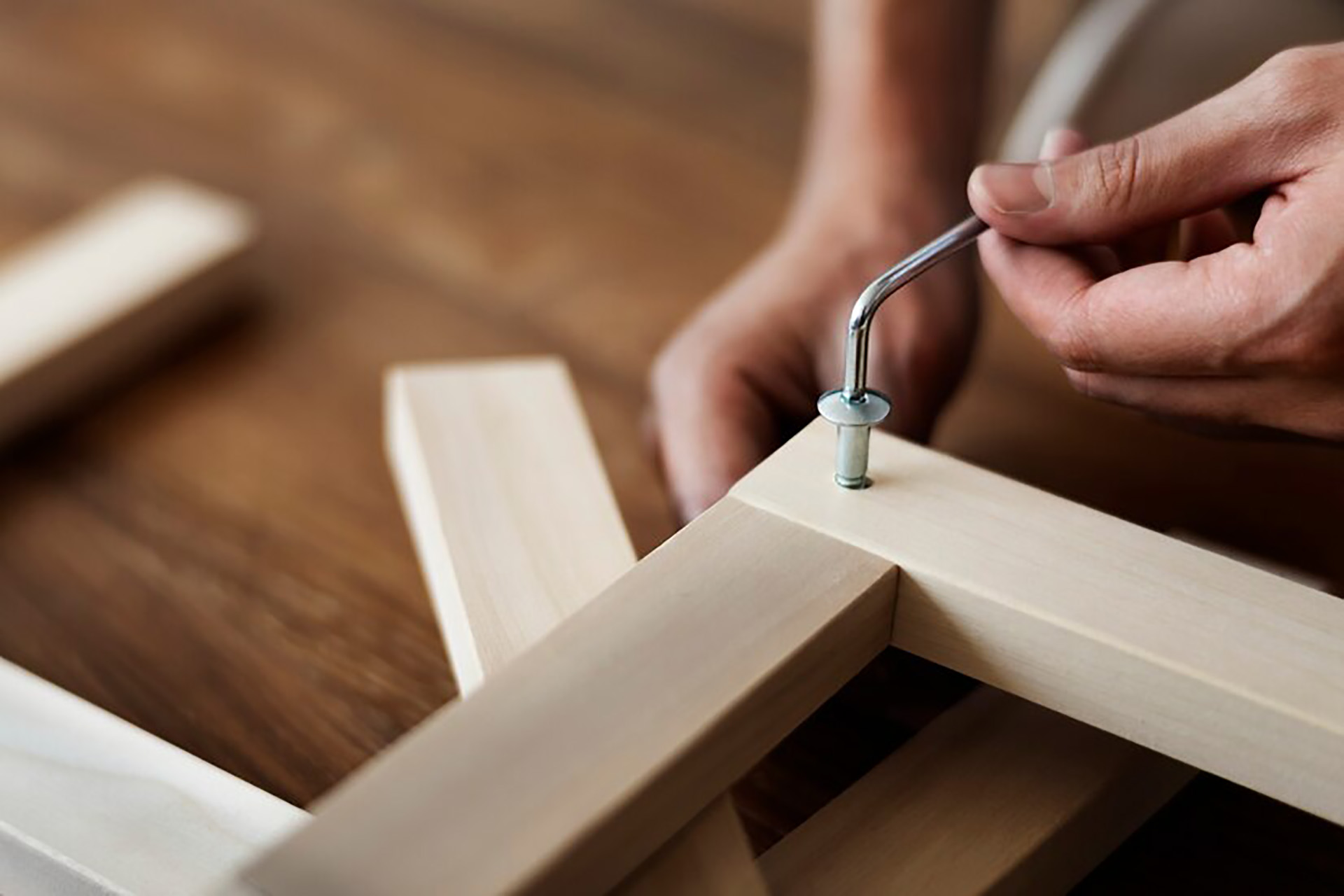 Hands using an Allen wrench to tighten a bolt into light-colored wood—furniture assembly in progress.